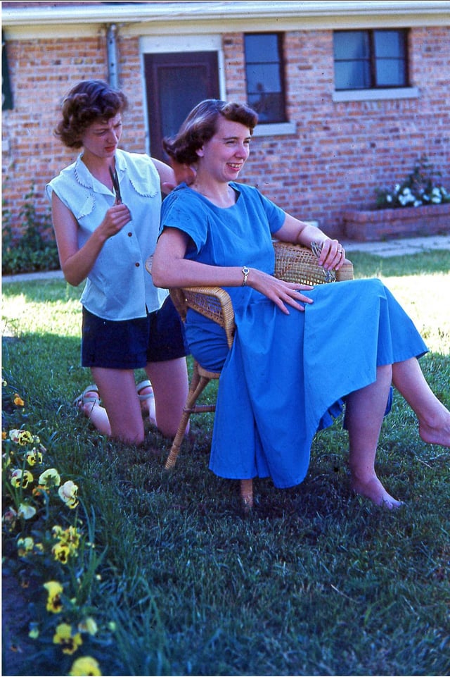 Young lady giving a hair cut to her frient outside her home, circa 1950s.