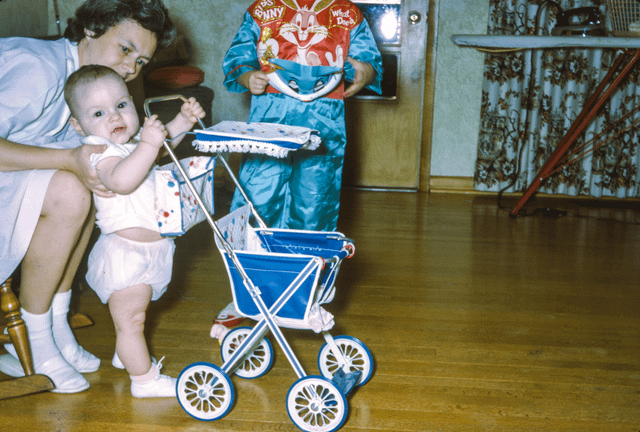 Mother poses her chubby baby besides a pram. Is a toy one or a real one? (seems small), October of 1950s.